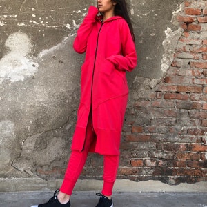 May include: A long, red hooded sweatshirt with a black zipper and a black drawstring. The sweatshirt has a long, flowing design and a relaxed fit. The model is wearing the sweatshirt with black sweatpants and black sneakers.
