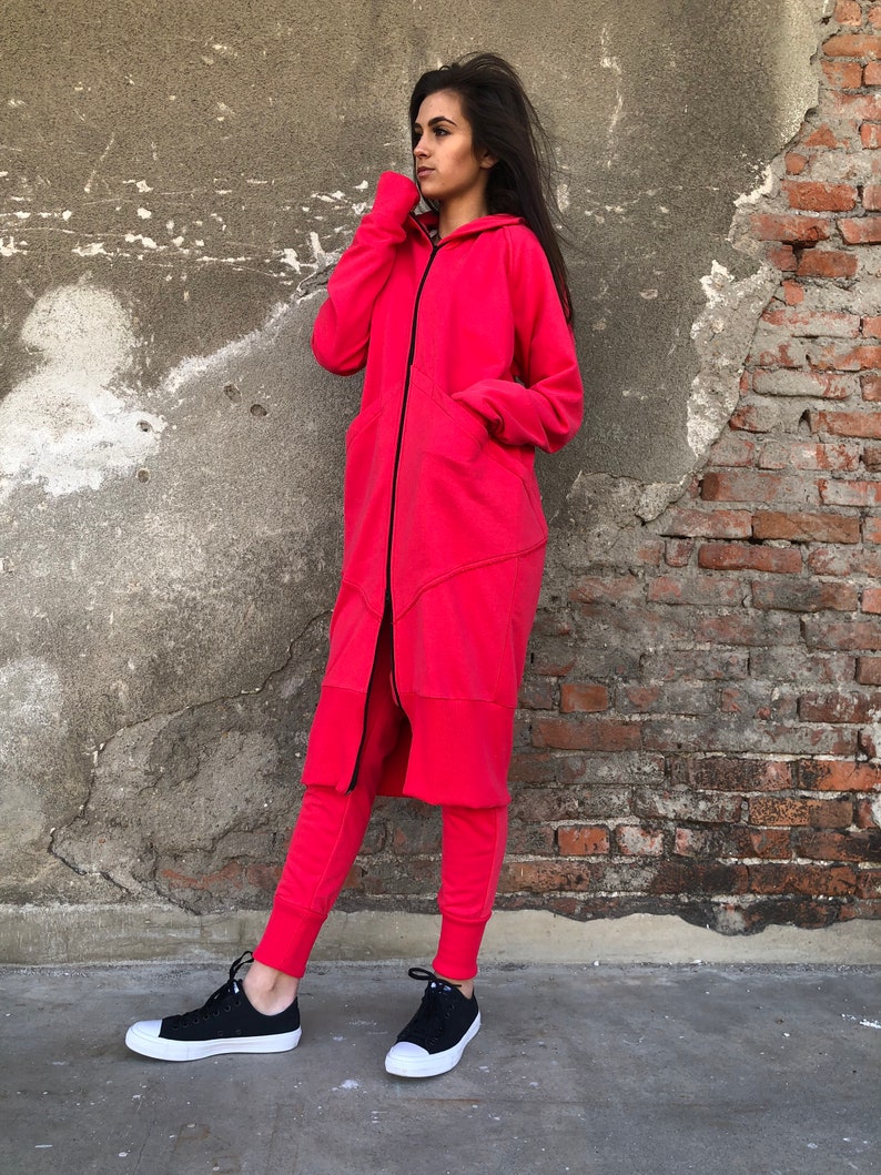 May include: A long, red hooded sweatshirt with a black zipper and a black drawstring. The sweatshirt has a long, flowing design and a relaxed fit. The model is wearing the sweatshirt with black sweatpants and black sneakers.