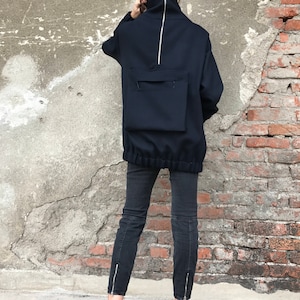 May include: A navy blue mock neck sweatshirt with a large zipper down the back and a large pocket on the back. The sweatshirt has a drawstring at the bottom. The person is wearing black jeans and pink sneakers.