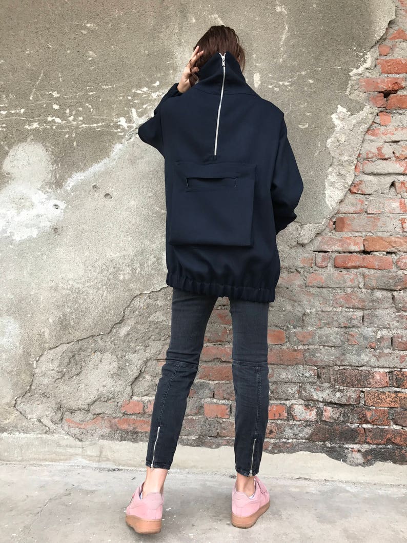 May include: A navy blue mock neck sweatshirt with a large zipper down the back and a large pocket on the back. The sweatshirt has a drawstring at the bottom. The person is wearing black jeans and pink sneakers.
