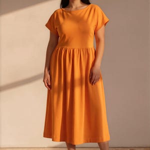 May include: A bright orange midi dress featuring a round neckline, short sleeves, and a gathered waistline that flows into a full, pleated skirt. The garment is paired with neutral-toned athletic sneakers, creating a simple and functional ensemble.