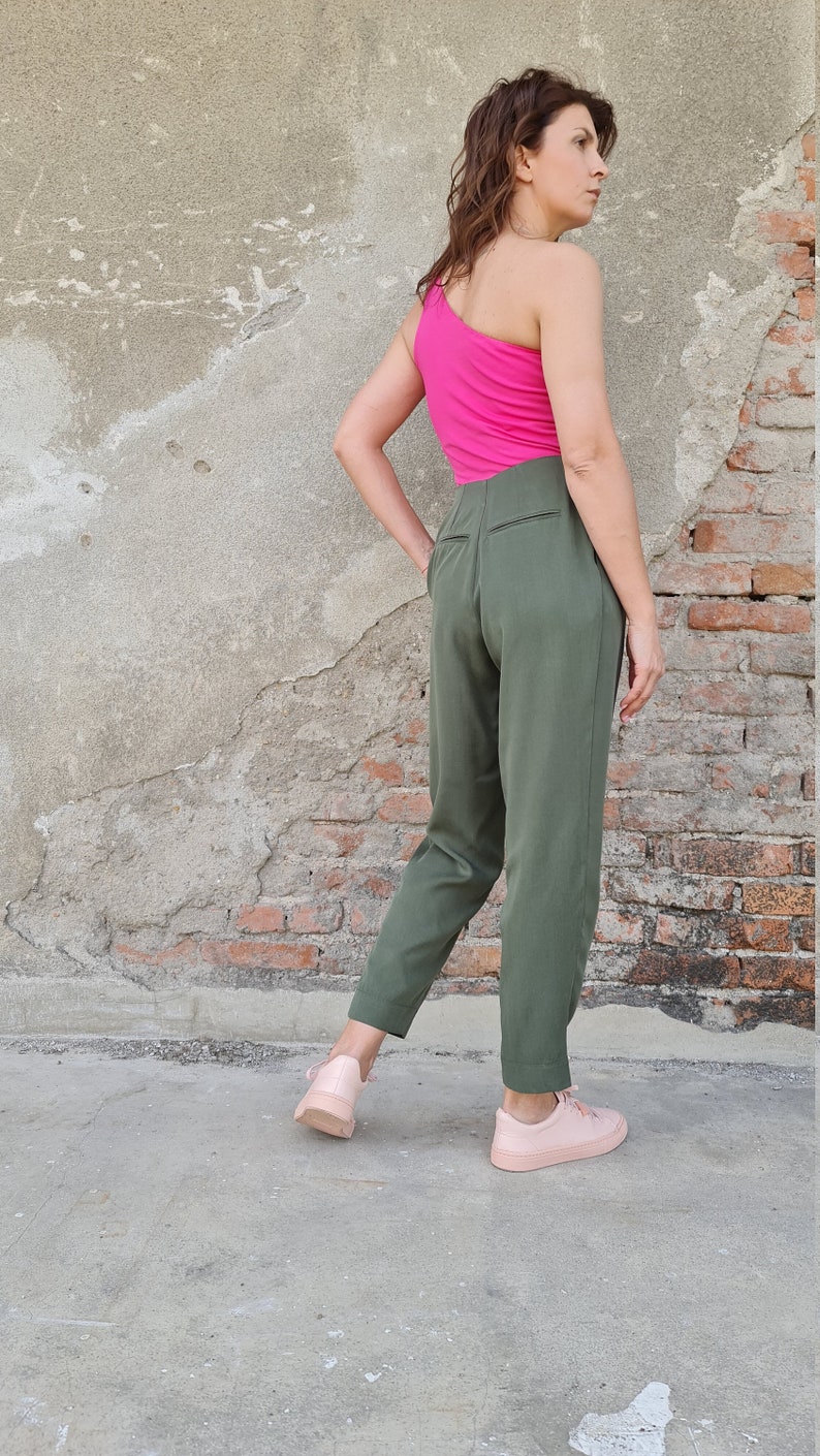 Puede incluir: Una mujer con un top rosa de un solo hombro y pantalones verde oliva. Est&aacute; de pie frente a una pared de ladrillo y lleva zapatillas rosas.