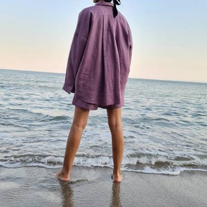 Może przedstawiać: Kobieta w jasnoliliowej koszuli i spodenkach z lnu stoi na piaszczystej plaży z oceanem w tle.