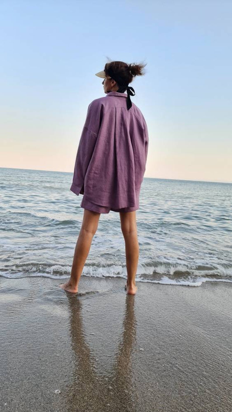 Może przedstawiać: Kobieta w jasnoliliowej koszuli i spodenkach z lnu stoi na piaszczystej plaży z oceanem w tle.