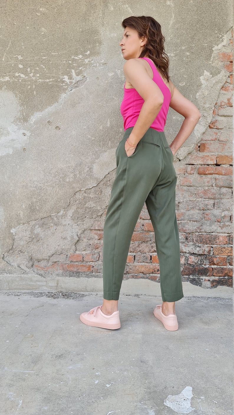 Puede incluir: Una mujer con un top rosa sin mangas y pantalones verde oliva de cintura alta. Lleva zapatillas rosas y est&aacute; de pie frente a una pared de ladrillo.
