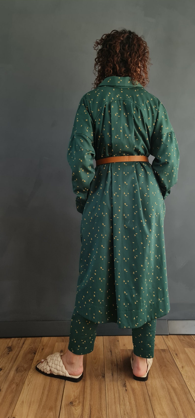 May include: A long green and yellow patterned shirt dress with a brown belt. The dress is worn with matching pants and cream colored sandals.