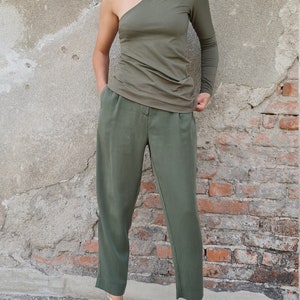 Puede incluir: Una mujer con un top verde oliva de un solo hombro con manga larga y pantalones verde oliva. Lleva zapatillas rosas.