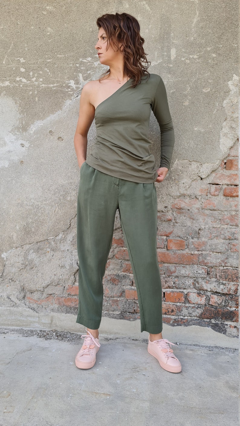 Puede incluir: Una mujer con un top verde oliva de un solo hombro con manga larga y pantalones verde oliva. Lleva zapatillas rosas.