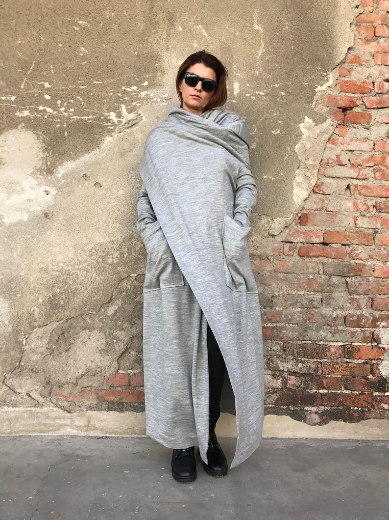May include: A long, gray, hooded cardigan with a draped design. The cardigan has two large pockets and a long, flowing hemline. The model is wearing black pants and black shoes.