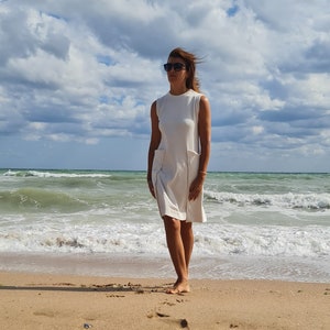May include: A woman wearing a white sleeveless dress and sunglasses stands on a sandy beach with ocean waves in the background.