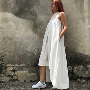 May include: A white sleeveless dress with a high neckline and a flowing skirt. The dress has a high-low hemline, with the front hemline ending above the knee and the back hemline trailing to the floor. The model is wearing white sneakers.