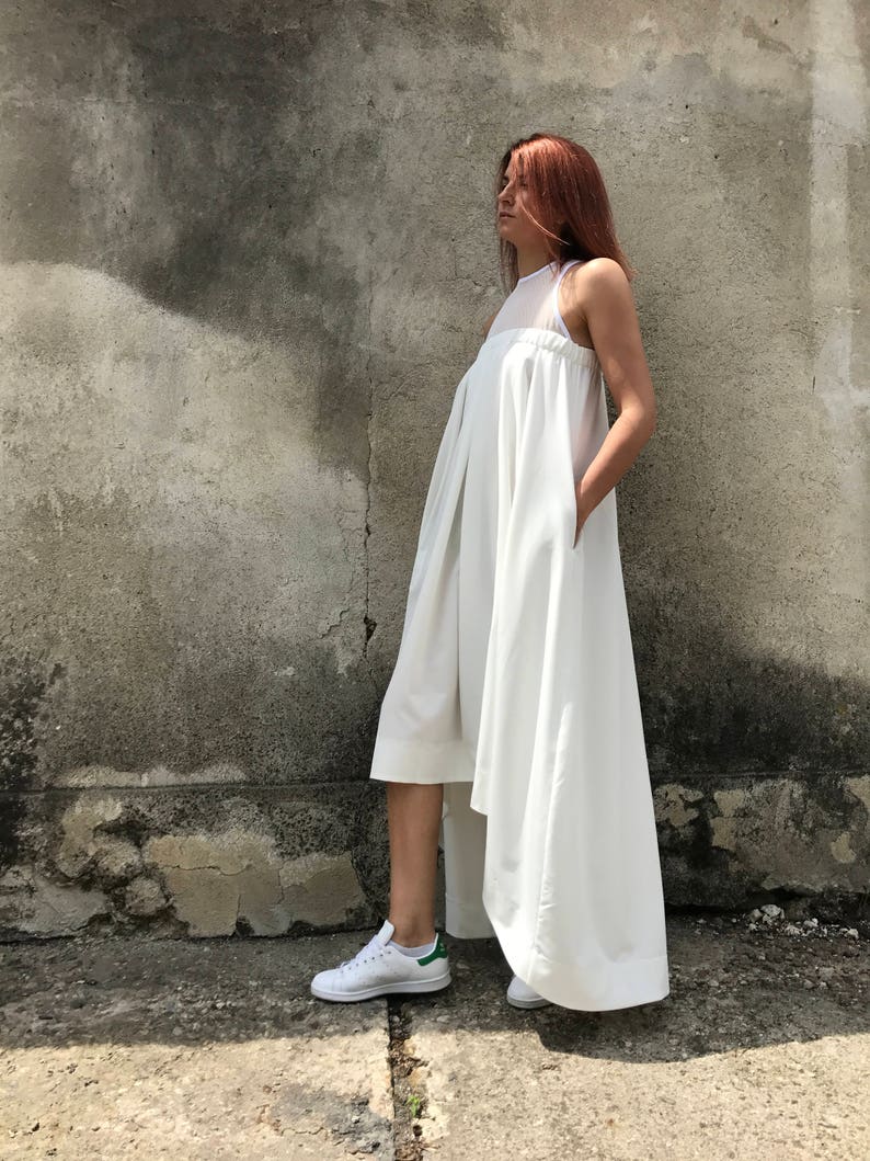 May include: A white sleeveless dress with a high neckline and a flowing skirt. The dress has a high-low hemline, with the front hemline ending above the knee and the back hemline trailing to the floor. The model is wearing white sneakers.