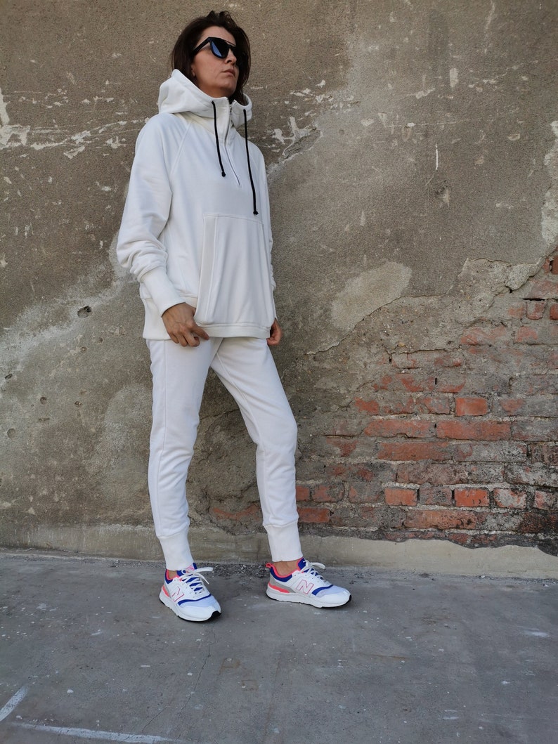 May include: A white hooded sweatshirt with a zipper and a kangaroo pocket. The model is wearing white sweatpants and white sneakers with blue and pink accents.