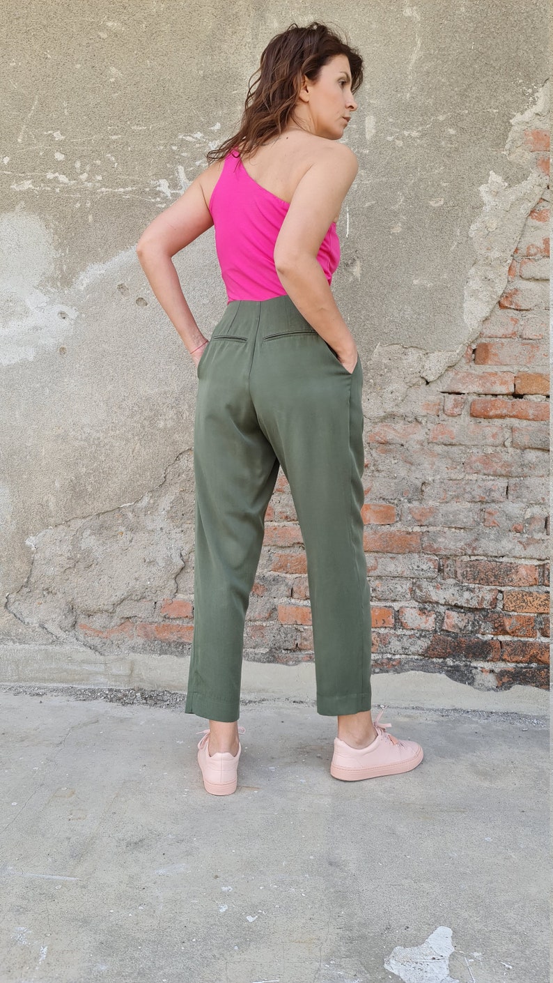 Puede incluir: Una mujer con un top rosa brillante de un solo hombro y pantalones verde oliva. Est&aacute; de pie con las manos en los bolsillos y mirando por encima del hombro. Lleva zapatillas de deporte rosa claro.