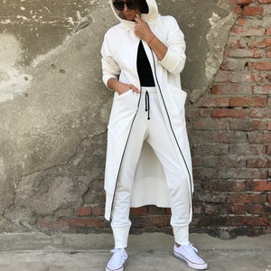 May include: A white hooded sweatshirt with a long, flowing design. The sweatshirt has a zipper closure and two large pockets. The model is wearing the sweatshirt with white pants and white sneakers.