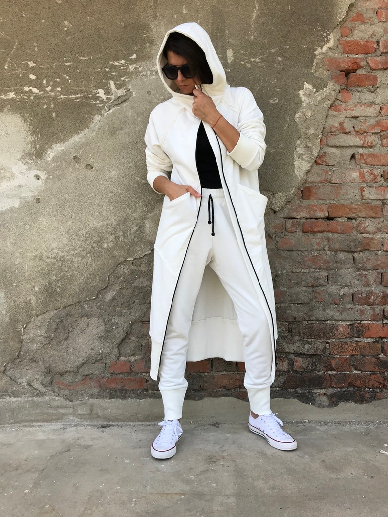 May include: A white hooded sweatshirt with a long, flowing design. The sweatshirt has a zipper closure and two large pockets. The model is wearing the sweatshirt with white pants and white sneakers.