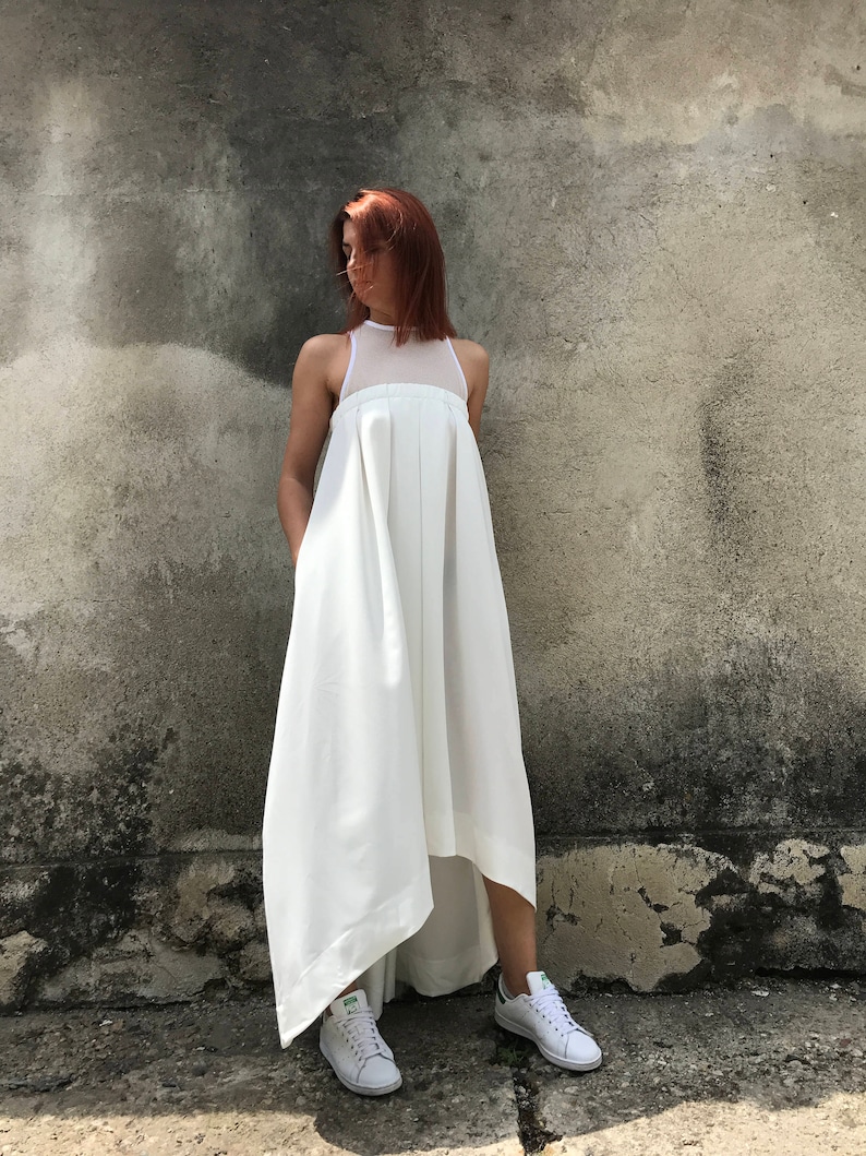 May include: A white maxi dress with a high neckline and a high-low hemline. The dress is made of a lightweight fabric and has a relaxed fit. The model is wearing white sneakers.