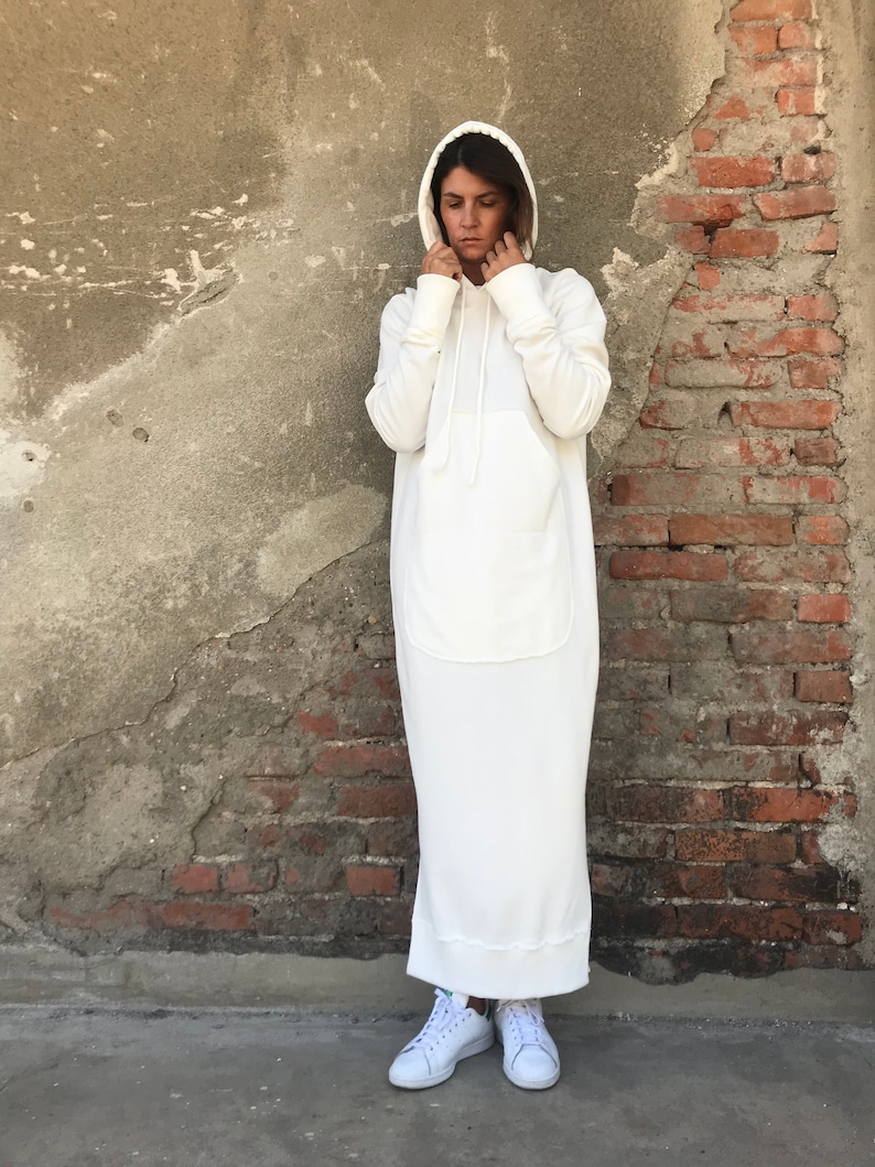 Pu&ograve; includere: Un abito bianco con cappuccio e una grande tasca sul davanti. L'abito &egrave; realizzato in un tessuto morbido e confortevole e ha una vestibilit&agrave; rilassata. La modella indossa sneakers bianche.