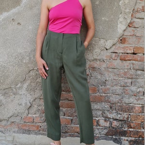 Puede incluir: Una mujer con un top rosa brillante de un solo hombro y pantalones verde oliva. Est&aacute; de pie frente a una pared de ladrillo y lleva zapatillas rosas.