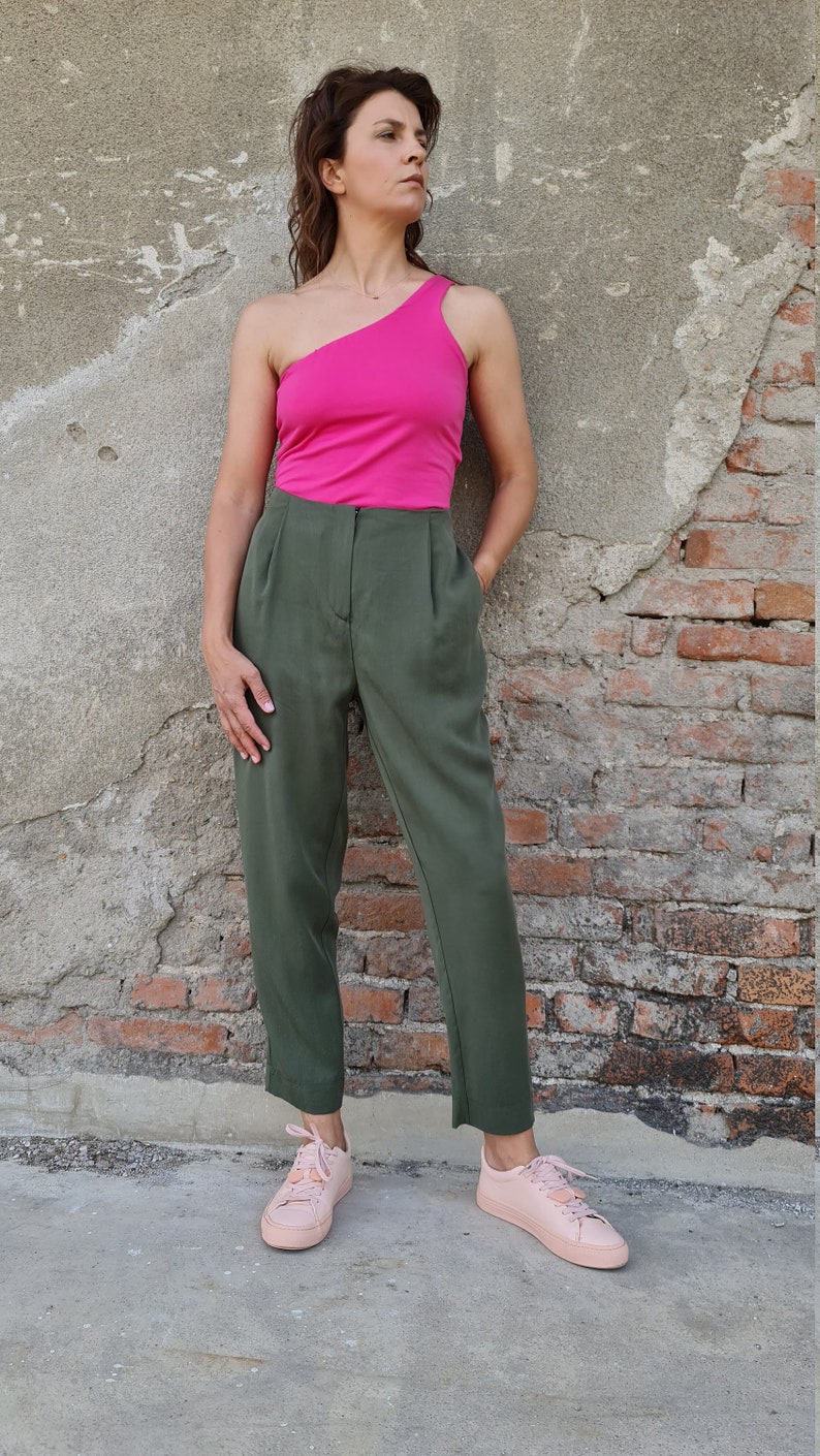 Puede incluir: Una mujer con un top rosa brillante de un solo hombro y pantalones verde oliva. Est&aacute; de pie frente a una pared de ladrillo y lleva zapatillas rosas.