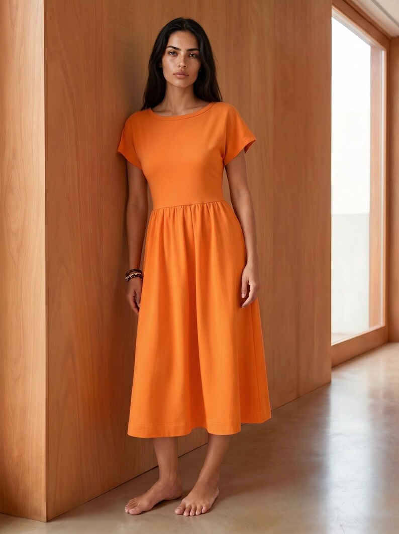May include: A solid orange midi dress featuring a round neckline, short sleeves, and a fitted bodice that transitions into a gathered, flowing skirt that falls below the knees. The garment is constructed from a smooth, opaque fabric.