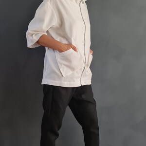 May include: A white zip-up jacket with a high collar and two large pockets. The jacket is worn over black pants and white sneakers.