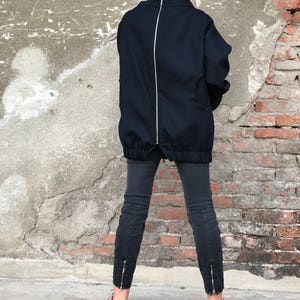 May include: A black oversized jacket with a full-length zipper down the back. The jacket has a high neck and a relaxed fit. The model is wearing black jeans and pink sneakers.
