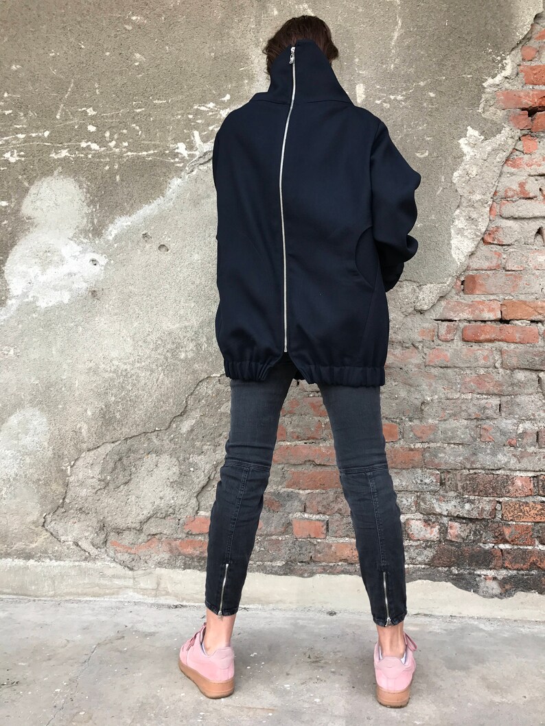 May include: A black oversized jacket with a full-length zipper down the back. The jacket has a high neck and a relaxed fit. The model is wearing black jeans and pink sneakers.