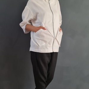May include: A white zip-up jacket with a high collar and two large pockets. The jacket is worn over black pants and white sneakers.