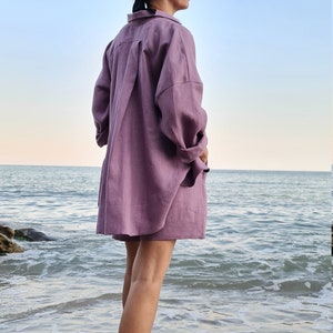 Może przedstawiać: Kobieta w komplecie koszuli i spodenek z lnu w kolorze lawendowym stoi na piaszczystej plaży z oceanem za sobą. Ma na sobie słomkowy daszek z czarną opaską.