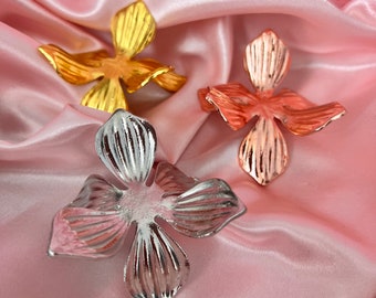 May include: Three metal flower embellishments in gold, rose gold, and silver. The flowers are lying on a pink satin fabric.