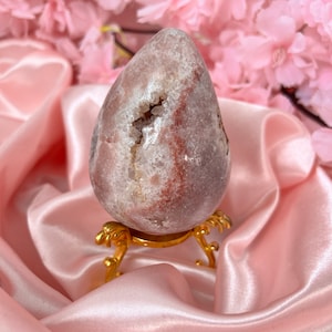 Könnte beinhalten: Ein rosa-weißes Agat-Geoden-Ei auf einem goldenen Metallständer. Die Geode hat eine natürliche Kristallbildung und ist ein beliebtes Dekorationsobjekt.