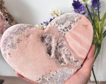 May include: A heart-shaped crystal geode in shades of pink and gray. The polished surface reveals a cavity filled with sparkling crystals. The geode is held in a hand, with a bouquet of flowers in the background.