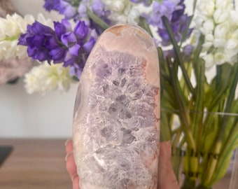 Puede incluir: Una gran geoda de amatista pulida con una formación de cristal natural y en bruto. La geoda es de color púrpura claro con un centro blanco translúcido. La geoda está sentada en una mano.