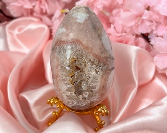 Puede incluir: Un huevo de cristal rosa y blanco con un centro de cuarzo transparente, sentado en un soporte de metal dorado. El huevo está sobre un fondo de tela de satén rosa.