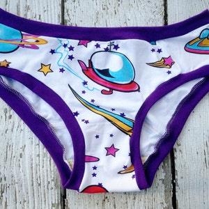 May include: White cotton underwear with a purple trim and a space-themed print featuring stars, planets, and a spaceship.