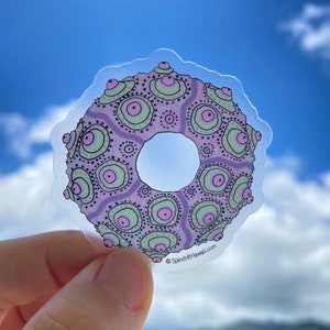 May include: A clear sticker with a purple, green, and pink patterned design of a sea urchin. The sticker has a hole in the center and the text "SpindriftHawaii.com" is printed on the bottom.
