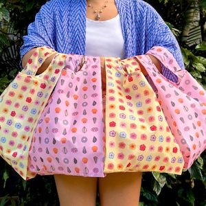 May include: A collection of reusable shopping bags in various colors, including yellow, pink, and lavender. Each bag features a repeating pattern of flowers and other small shapes. The bags have handles for easy carrying and are held in front of a person.