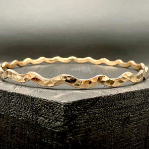 Könnte beinhalten: Ein goldfarbenes Armreif-Armband mit einem strukturierten, wellenförmigen Design. Das Armband hat eine gehämmerten Oberfläche, die ein facettiertes Aussehen erzeugt. Der Schmuck wird auf einer dunklen Oberfläche präsentiert.