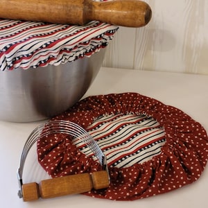 May include: A wooden rolling pin rests on a bowl covered with a red, white, and blue striped fabric. A red fabric bowl cover with white stars and a stainless steel pastry blender with a wooden handle are also present.