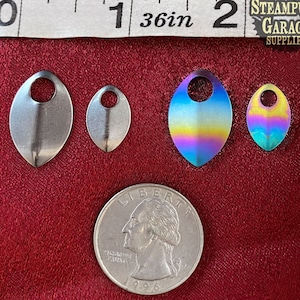 Könnte beinhalten: Vier Metall-Anhänger in verschiedenen Größen und Ausführungen. Zwei sind silberfarben, einer ist regenbogenfarben schillernd und einer ist mehrfarbig schillernd. Die Anhänger sind tropfenförmig mit einem Loch oben. Die Anhänger werden auf einer roten Oberfläche mit einer US-amerikanischen 25-Cent-Münze zur Größenbestimmung gezeigt.