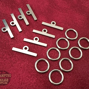 May include: A set of silver metal hardware for crafting, including 5 small bars with a loop at the end and 7 silver jump rings.  Steampunk Garage Supplies is printed on the red background.