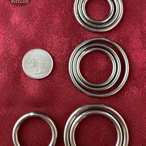 May include: Four sets of metal rings in different sizes, with the smallest ring being about the size of a US quarter. The rings are all silver in color and have a smooth, polished finish. The rings are arranged in a row on a red fabric background.
