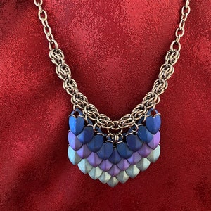 May include: A silver chain necklace with a pendant made of multiple heart-shaped metal scales in shades of purple, blue, and silver.