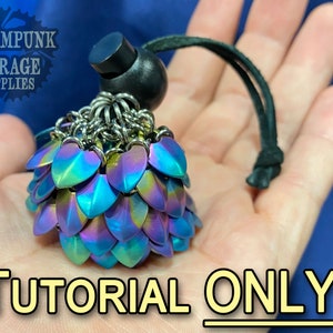 May include: A close-up of a small, iridescent blue, green, and purple chainmaille pendant with a black cord. The pendant is in the palm of a hand. The text "TUTORIAL ONLY!" is below the pendant.