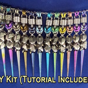 May include: A collection of colorful, handmade metal pendants hanging from a chain. Each pendant features a series of silver-colored metal scales and a long, pointed spike in various iridescent colors. The text "DIY KIT (TUTORIAL INCLUDED!)" is at the bottom.