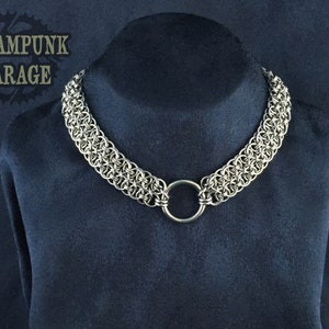 May include: A silver chainmail necklace with a large silver ring. The necklace is on a dark blue velvet background.