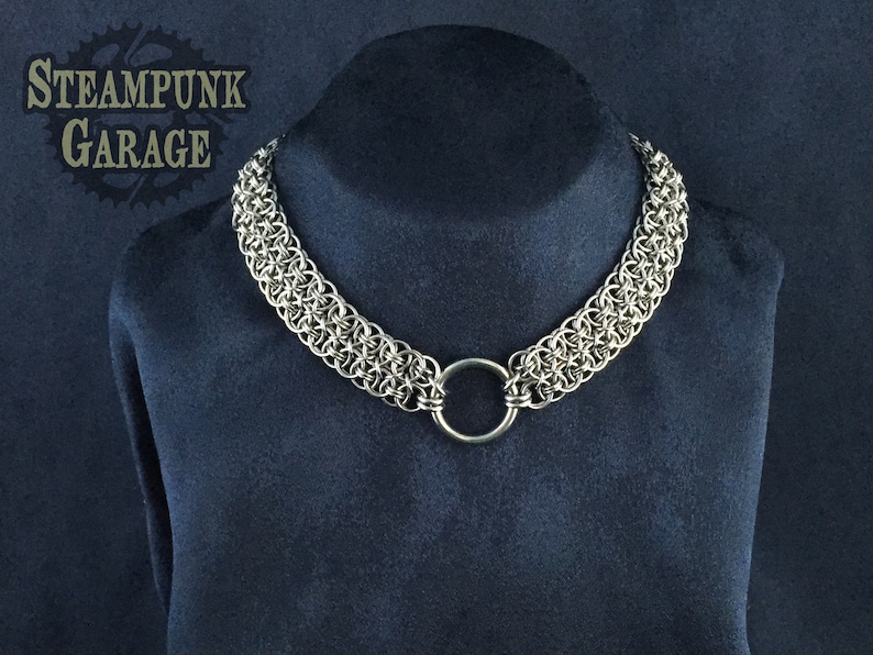 May include: A silver chainmail necklace with a large silver ring. The necklace is on a dark blue velvet background.