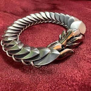 May include: A silver-toned bracelet with a dragon head clasp. The bracelet is made of overlapping, shield-shaped metal pieces linked together. The dragon head features intricate details and is positioned at the end of the bracelet. The bracelet is displayed on a red surface.