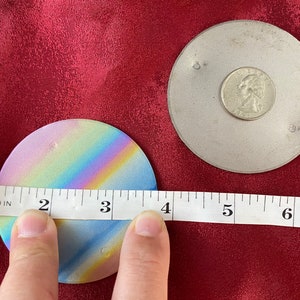 May include: Two round coasters. One coaster has a rainbow gradient design and measures approximately 10 cm in diameter. The other coaster is silver and has a coin on top. The coasters are on a red surface.